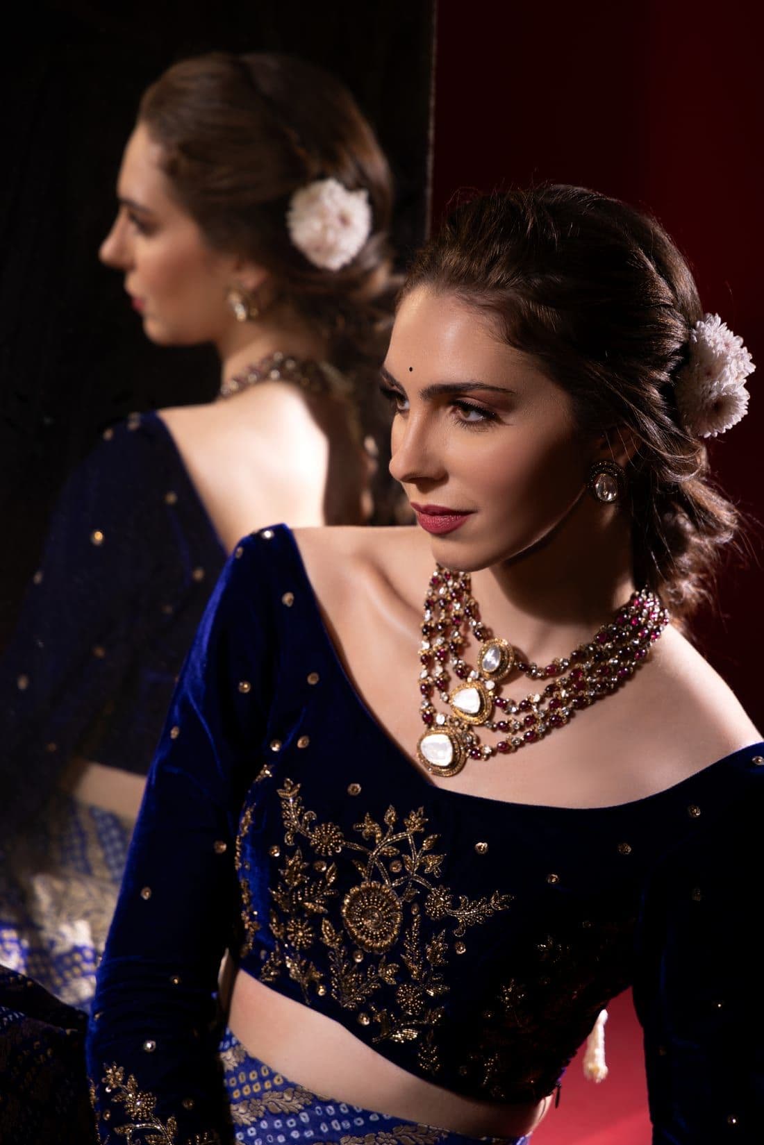 Woman in a navy velvet blouse and layered ruby necklace beside a mirror