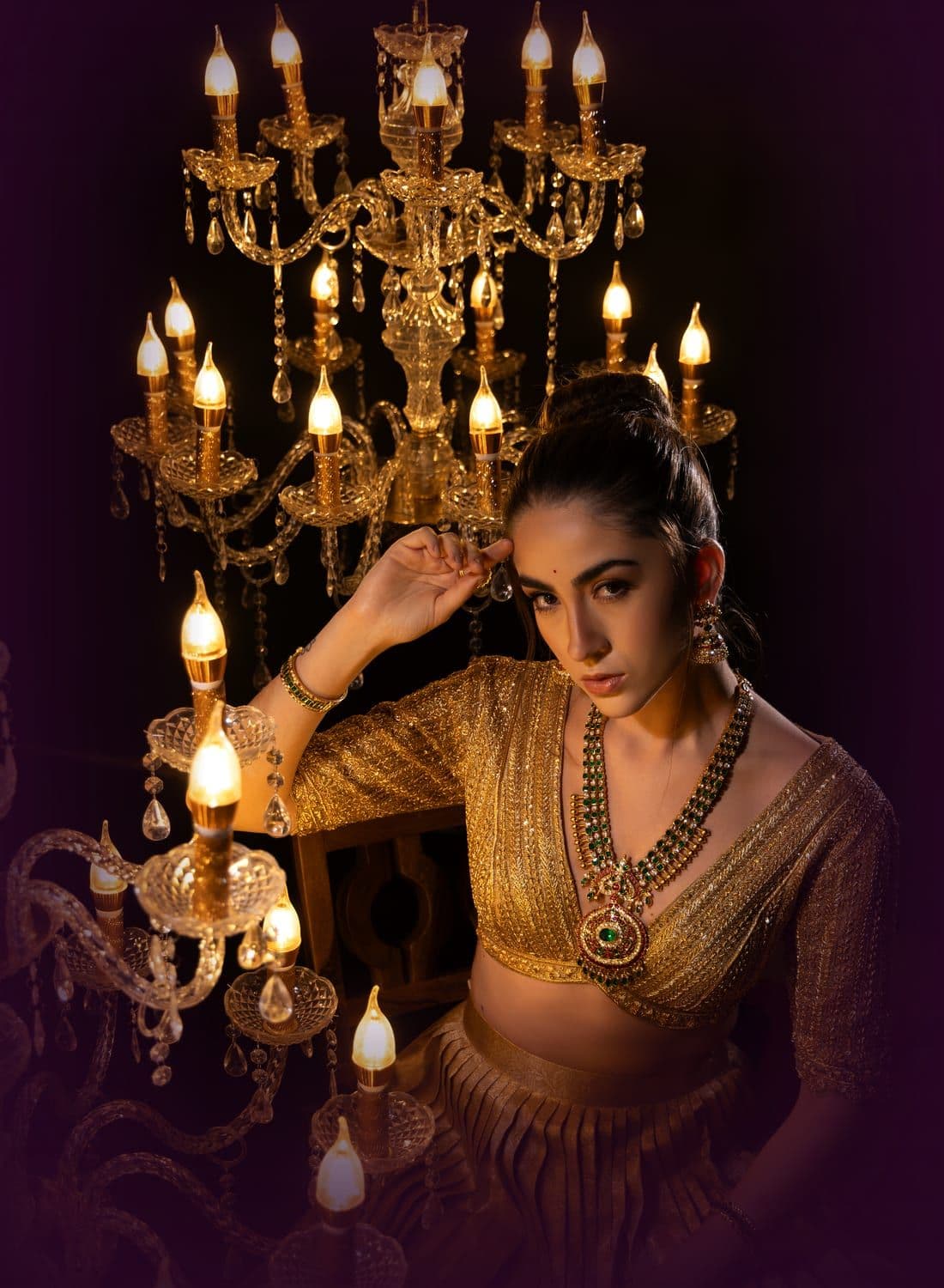 Woman in a gold lehenga seated among lit crystal chandeliers