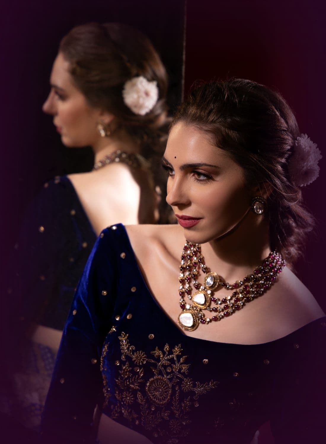 Woman in a navy velvet blouse and layered ruby necklace beside a mirror